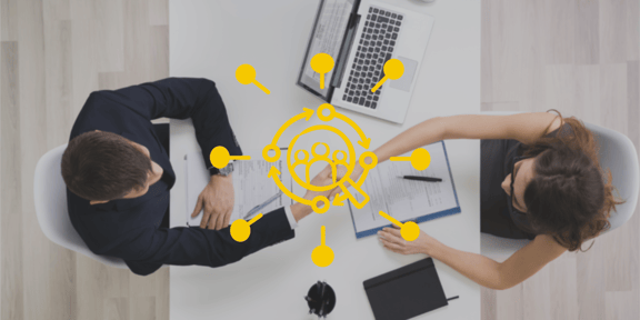 Ein Foto aus der Vogelperspektive: zwei Personen sitzen an einem Tisch und geben sich die Hand. Im Vordergrund ein Icon: eine Lupe die auf Personen zeigt, drum herum Punkte, die davon weg gehen.