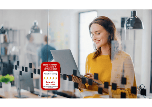 : Junge Frau steht lächelnd mit Laptop in der Hand hinter einer Glaswand – Symbolbild für ein modernes Recruitingmanagement