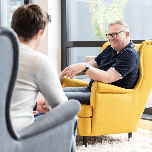zwei Personen sitzen gegenüber voneinander in zwei Sesseln. Einer ist grau, der andere ist gelb. Sie lachen miteinander