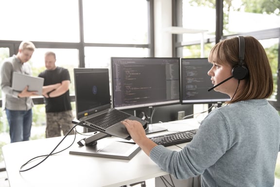 A young woman with a headset sits in front of a laptop and two screens displaying programming code - a symbol of beesite's service quality.