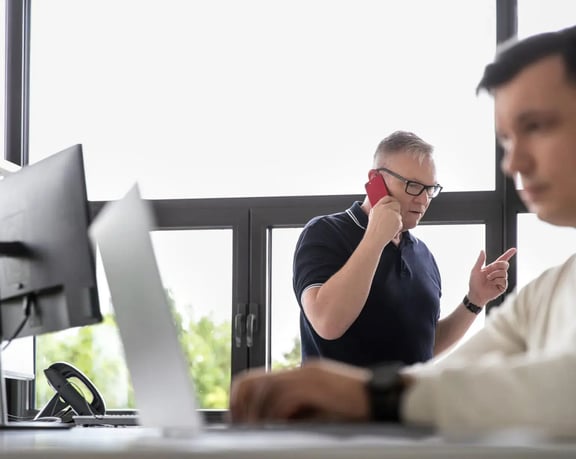 A man on the phone gestures with his hands while another man sits at a laptop in the blurred foreground - a symbolic image for a recruitment solutions consultancy.