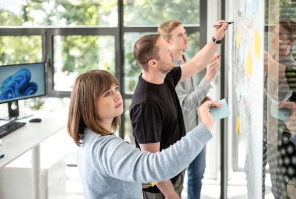 A young woman and two men write on post-its stuck to a glass wall - a symbolic image for collective brainstorming and innovative idea generation.
