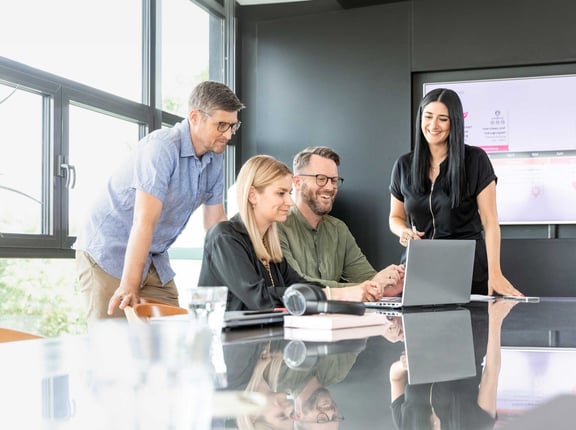 Four people in a modern meeting room collaborating at a laptop
